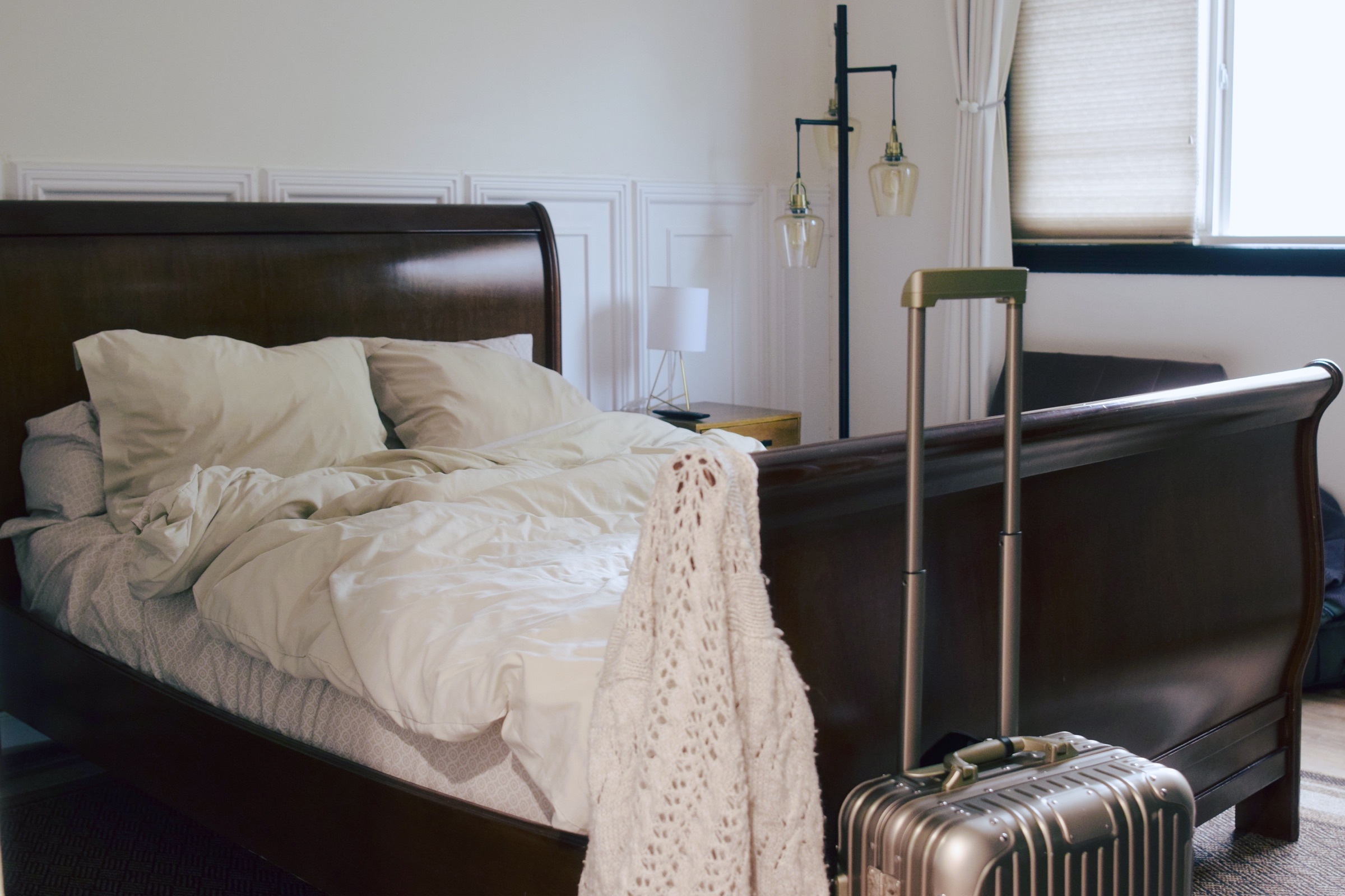 Rumpled white bedding on a dark sleigh bed with a gold suitcase and knit cardigan beside it