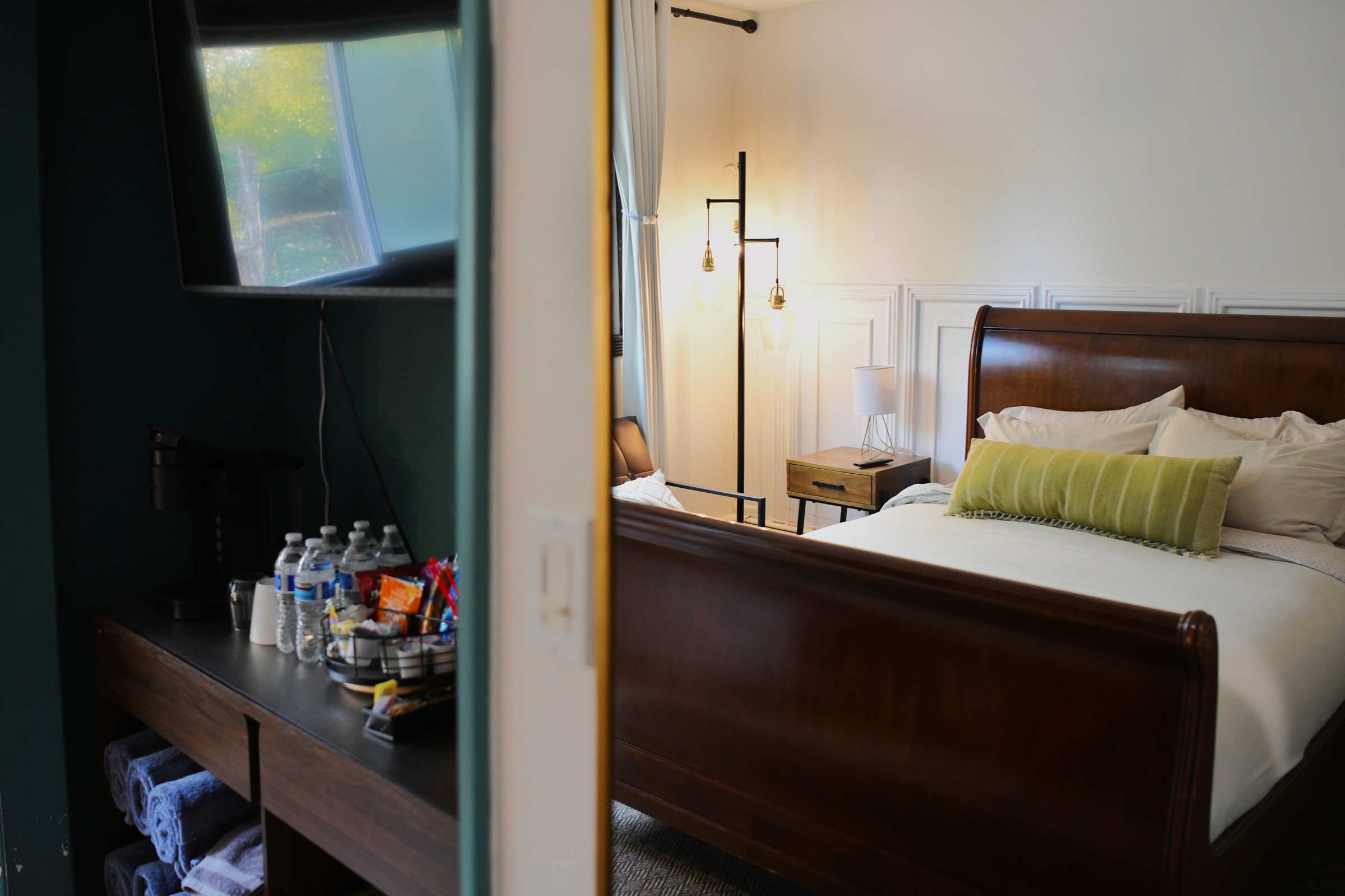 A bed lit by a floor lamp, with the coffee and snack station reflected in a mirror