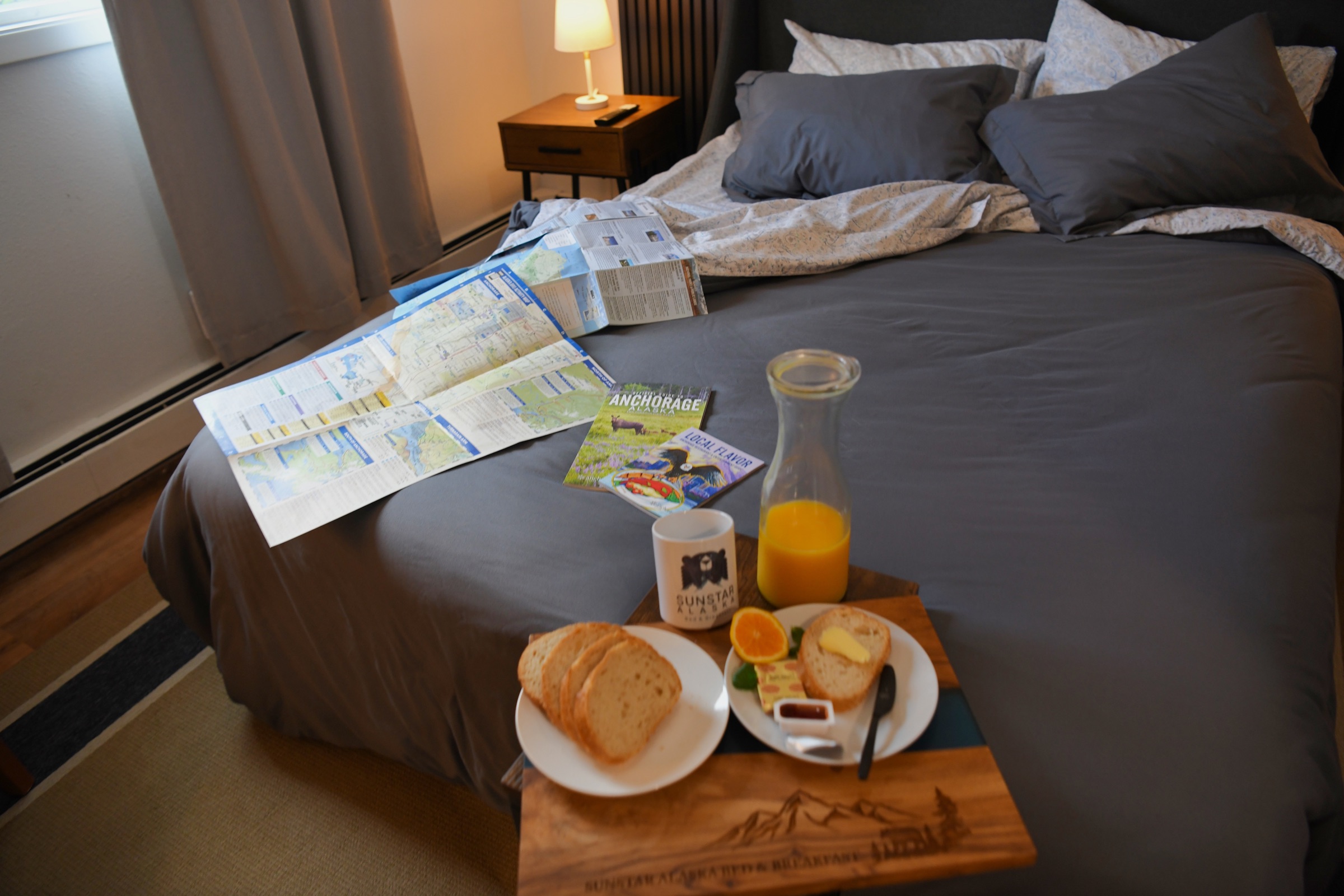 A breakfast tray with toast, juice and a SunStar mug on a grey duvet, with Anchorage maps beside it