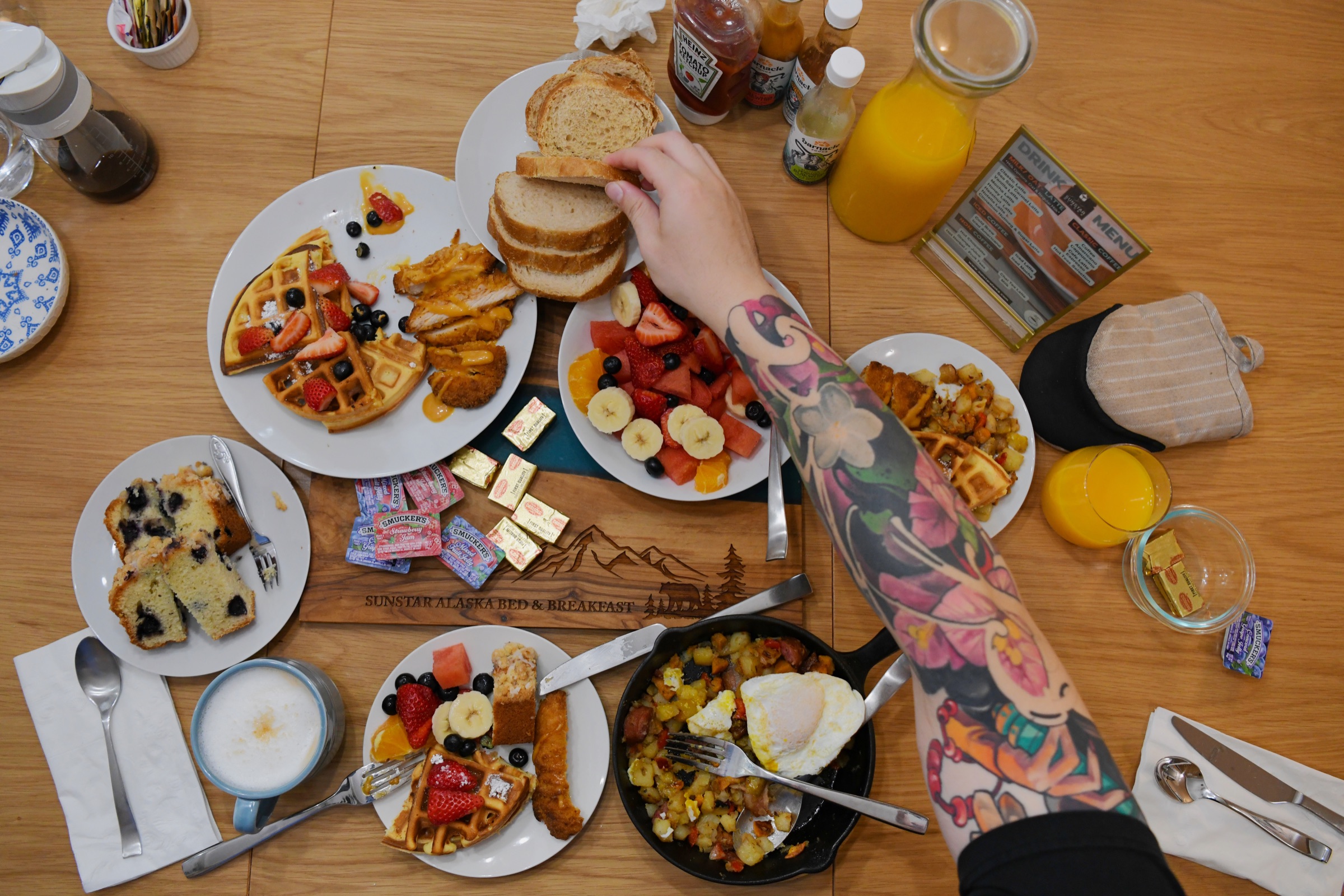 An overhead view of a breakfast table of waffles, fruit, hash and juice with a tattooed arm reaching for toast